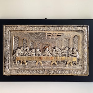 Può includere: Una placca da parete rettangolare che raffigura L'Ultima Cena con una finitura metallica argento e oro. La scena in rilievo è racchiusa in una semplice cornice nera con un bordo decorativo caratterizzato da viti e foglie in rilievo.