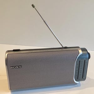 Könnte beinhalten: Ein silbernes und schwarzes tragbares Sony-Radio mit ausziehbarer Antenne. Das Radio hat eine Lautsprecherabdeckung aus Mesh und ein Einstellrad an der Seite. Das Sony-Logo ist auf der Vorderseite sichtbar.