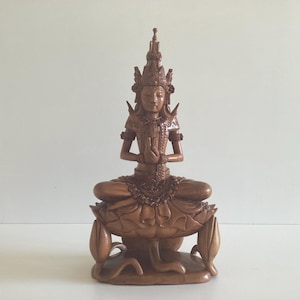 May include: A detailed wooden statue of a seated figure in a meditative pose. The figure wears an ornate headdress and is seated on a lotus flower base. The statue is carved from a light brown wood and has intricate details.