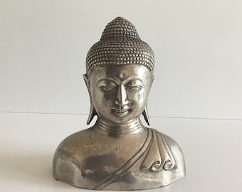 May include: A silver-colored bust of Buddha with a serene expression. The sculpture features a detailed, textured hairstyle and draped robes. The bust is a decorative item, likely for home decor or spiritual practice.