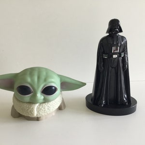 May include: A green Grogu figure and a black Darth Vader figurine. The Grogu figure has large black eyes and large ears. The Darth Vader figurine is in a classic pose, with a black cape and helmet.