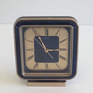 Puede incluir: Reloj cuadrado Orient con esfera dorada y borde azul marino. El reloj presenta agujas doradas y números romanos. La base y el marco son dorados. El reloj es de estilo vintage y se encuentra sobre una superficie blanca.