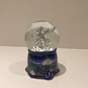 Könnte beinhalten: Eine Schneekugel mit einem weißen geflügelten Pferd in einer klaren Glaskugel. Der blaue Sockel hat eine Nachtszene und eine Schlossillustration. Die Kugel ist ein dekorativer Artikel.