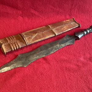 May include: An antique sword with a dark, weathered blade and a carved handle, lying next to its leather sheath. The sheath is a light brown color with decorative stitching and geometric patterns. The items are set against a red background.
