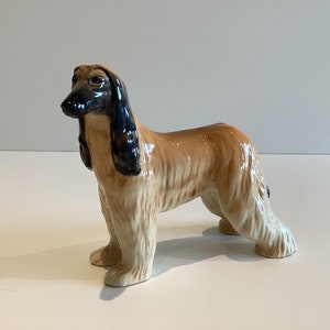Puede incluir: Figura de cerámica de un galgo afgano. El perro está representado de pie, con un cuerpo largo y esbelto y un pelaje fluido. La figura presenta una paleta de colores beige, negro y crema, con un acabado brillante. La cabeza del perro está ligeramente girada hacia un lado.