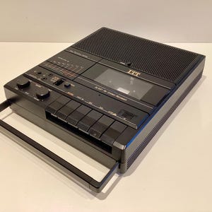 Könnte beinhalten: Schwarzer ITT Stereo-Kassettenrekorder mit eingebautem Lautsprecher und Griff. Der Rekorder hat Knöpfe, Drehregler und ein Kassettenfach. Die Modellnummer ist auf der Vorderseite sichtbar. Das Gerät steht auf einer weißen Oberfläche.