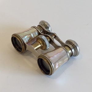 May include: Vintage opera glasses with a mother-of-pearl finish. The binoculars have a silver frame with gold accents. The lenses are black. The focus wheel is gold. A classic item.