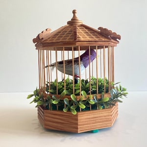May include: A decorative wooden birdcage with a small bird inside. The cage is made of light brown wood and has a hexagonal base. The bird has purple and gray feathers and is perched on a bed of green leaves.