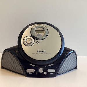 Op de afbeelding: Een draagbare Philips cd-speler in zilver en marineblauw. De speler heeft een ronde bovenkant met een digitaal display en bedieningsknoppen. De basis heeft twee luidsprekerroosters en bedieningsknoppen.