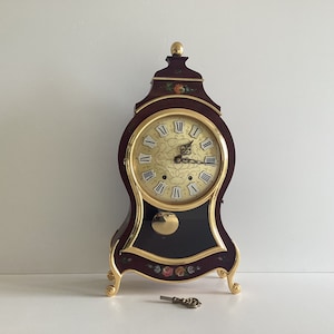 May include: A decorative wooden clock with a dark burgundy finish and gold accents. The clock face is gold with black Roman numerals. Floral designs adorn the clock's face and base. A small key rests in front of the clock.