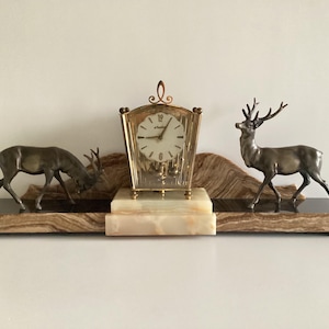 May include: A decorative mantel clock featuring a gold-toned clock with a white face, flanked by two bronze-colored deer figurines. The clock and deer are set on a black and beige marble base, creating a classic and elegant display.