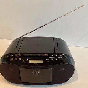 Könnte beinhalten: Schwarzer Sony-Boombox mit silberner Antenne. Die Boombox verfügt über einen CD-Player, einen Kassettenrekorder und ein Radio. Auf der Vorderseite befinden sich ein Lautsprecher und das Sony-Logo. Es hat ein digitales Display und Tasten zur Audiosteuerung.