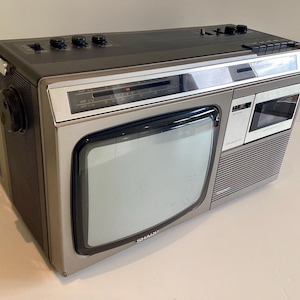 May include: Vintage Sharp portable TV with a built-in cassette player and radio. The unit has a silver and grey colour scheme with black control knobs. The screen is a square shape. The unit has a handle for easy carrying.