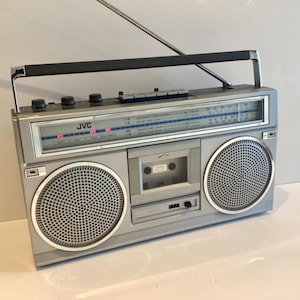 Op de afbeelding: Zilveren JVC-boombox met een zwarte handgreep en antenne. De radio heeft twee grote luidsprekerroosters, een cassettespeler en een radioschaal. Het JVC-logo is zichtbaar. Een klassiek retro audio-apparaat.