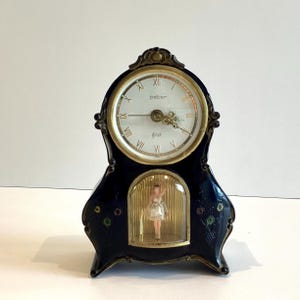 Könnte beinhalten: Eine marineblaue Kaminuhr mit einem goldfarbenen Zifferblatt und Zeigern. Das Zifferblatt hat römische Ziffern und das Wort "peter". Unter dem Zifferblatt ist eine kleine Ballerina-Figur in einem Glaskasten zu sehen. Die Uhr ist mit farbigen Akzenten verziert.