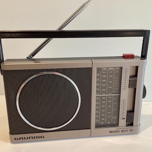 May include: Vintage Grundig Music Boy 60 portable radio. The radio has a silver and black color scheme with a mesh speaker cover and a silver circular accent. It features a telescoping antenna and a carrying handle.