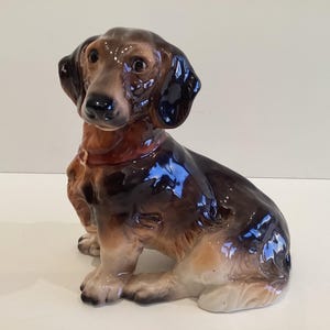 Puede incluir: Figura de perro salchicha de cerámica en posición sentada. El perro tiene un acabado brillante con colores marrón, negro y canela. La figura tiene detalles, incluyendo una cara y patas realistas.