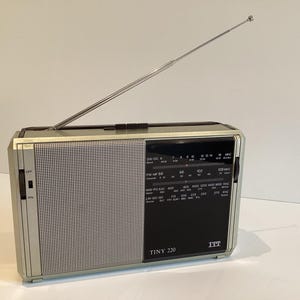 May include: A silver and black vintage radio with an extendable aerial. The radio features a speaker grille and a black control panel with the text "TINY 220" and "ITT". It sits on a white surface.
