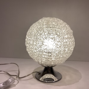 May include: A spherical table lamp with a textured, woven-like shade. The lamp has a chrome base and a clear cord with an on/off switch. The light shines brightly through the shade.