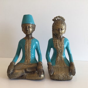 Peut inclure: Deux statues assises de personnages en tenue traditionnelle. Les figures ont la peau couleur bronze et sont ornées de vêtements turquoise et dorés. L'une porte un chapeau, tandis que l'autre a une coiffe élaborée. Les statues sont probablement des objets décoratifs.