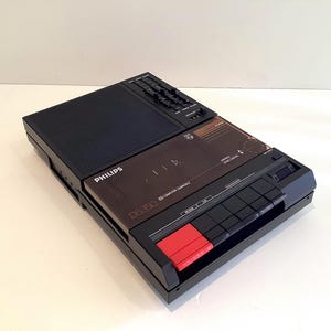 Puede incluir: Una grabadora de casete Philips D6350 negra con una rejilla de altavoz rectangular, una puerta transparente para el compartimento de casetes y un panel de control con botones negros y un botón rojo destacado para funciones de grabación.