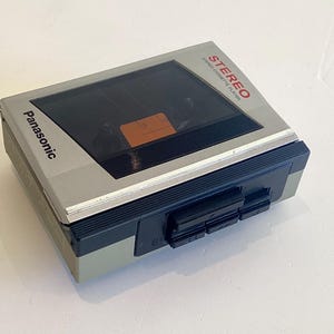 May include: A vintage Panasonic stereo cassette player in a silver and black case. The player has a clear window to view the cassette tape and a series of black buttons. The word "STEREO" is printed in red.