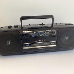 Könnte beinhalten: Ein schwarzer, tragbarer Panasonic Stereo-Kassettenrekorder mit einem Tragegriff an der Oberseite, zwei seitlichen Lautsprechern, einem zentralen Radiotuner mit Frequenzskalen und einem Kassettenfach mit der Aufschrift Full Auto Stop und Cue & Review.