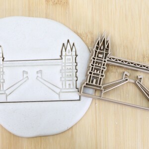 Może przedstawiać: Metalowa foremka do ciasteczek w kształcie Tower Bridge w Londynie. Foremka jest dopuszczona do kontaktu z żywnością i jest pokazana obok białego odcisku mostu w fondancie.