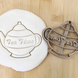 Könnte beinhalten: Ein brauner Plastik-Ausstecher in Form einer Teekanne mit der Aufschrift "Tea Time". Der Ausstecher liegt auf einer Holzoberfläche neben einem weißen Teigabdruck des Ausstechers.