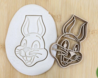 May include: A brown wooden cookie cutter in the shape of Bugs Bunny's head. The cutter is shown next to a white imprint of the cutter in a block of modeling clay.