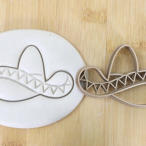 Könnte beinhalten: Ein brauner Plastik-Ausstecher in Form eines Sombreros. Der Ausstecher liegt auf einer Holzoberfläche neben einem Stück weißen Fondants mit einer Abformung des Sombreros.