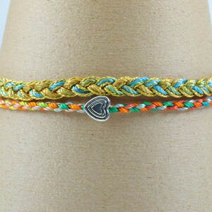Könnte beinhalten: Zwei geflochtene Armbänder. Das obere Armband ist goldfarben mit blauen und hellblauen Akzenten. Das untere Armband ist mehrfarbig mit einem silbernen Herzanhänger. Die Armbänder werden auf einem braunen, kegelförmigen Objekt präsentiert.