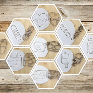 Könnte beinhalten: Ein Set mit 10 Ausstechern in Form von medizinischen Symbolen, darunter ein Herz mit einer Herzschlaglinie, ein Verband, eine Pille, ein Erste-Hilfe-Kasten, eine Krankenschwestermütze, eine Spritze, ein Fläschchen, ein Blutbeutel und ein Krankenwagen. Die Ausstecher sind aus weißem Kunststoff und in einem sechseckigen Muster auf einem Holzgrund angeordnet.