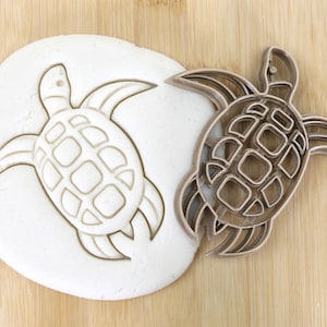 Puede incluir: Un cortador de galletas de plástico marrón en forma de tortuga marina. El cortador está sobre una superficie de madera junto a una masa con la forma de tortuga presionada en ella. El cortador es de calidad alimentaria.