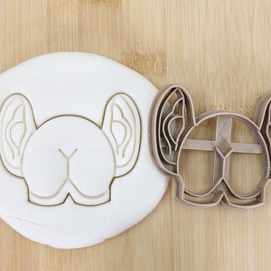 Puede incluir: Un cortador de galletas de plástico marrón en forma de cabeza de perro con orejas grandes. El cortador está sobre una superficie de madera junto a una pieza de masa blanca con la misma forma recortada.