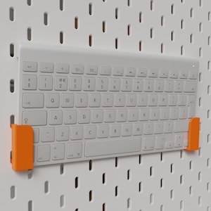 Könnte beinhalten: Eine weiße drahtlose Tastatur mit orangefarbenen Clips an den Seiten. Die Tastatur hängt an einer weißen Lochwand.