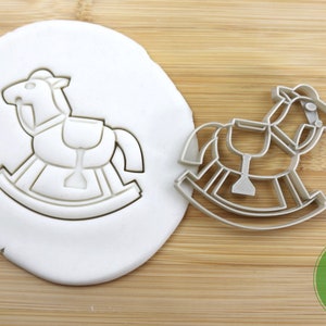 Puede incluir: Un cortador de galletas de caballo de balanc&iacute;n plateado sobre una superficie de madera. El cortador se muestra presionado en un fondant blanco. El cortador es de calidad alimentaria.