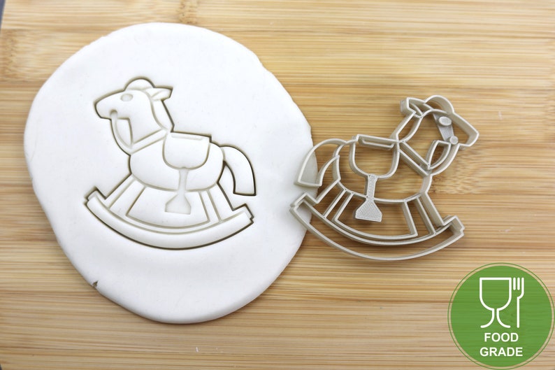 Puede incluir: Un cortador de galletas de caballo de balanc&iacute;n plateado sobre una superficie de madera. El cortador se muestra presionado en un fondant blanco. El cortador es de calidad alimentaria.