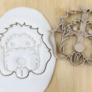 May include: A brown plastic cookie cutter in the shape of a dog's head. The cutter is on a wooden surface next to a piece of white dough with the dog's head cut out.