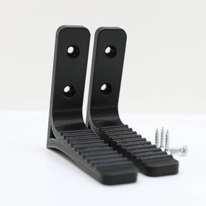 Puede incluir: Dos soportes de pared de plástico negro con una superficie texturizada y cuatro agujeros para tornillos. Los soportes están diseñados para sujetar objetos en una pared. Se incluyen cuatro tornillos plateados.