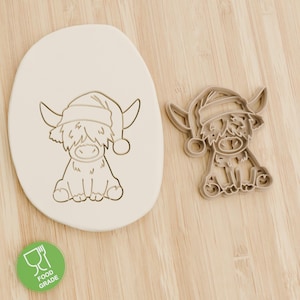 May include: A Highland cow cookie cutter and a pressed cookie, both featuring the same design. The cow is wearing a Santa hat. The cookie cutter is made of plastic. A green food grade sticker is in the lower left corner.