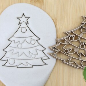 Puede incluir: Un cortador de galletas de &aacute;rbol de Navidad de pl&aacute;stico de calidad alimentaria. El cortador se muestra sobre una superficie de madera con un trozo de arcilla blanca detr&aacute;s. El cortador est&aacute; dise&ntilde;ado para crear una galleta de &aacute;rbol de Navidad con una estrella en la parte superior y adornos.