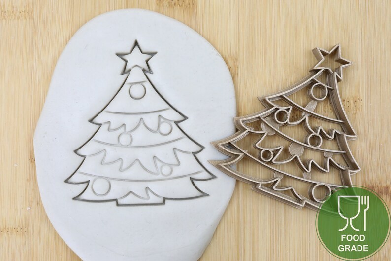 Puede incluir: Un cortador de galletas de &aacute;rbol de Navidad de pl&aacute;stico de calidad alimentaria. El cortador se muestra sobre una superficie de madera con un trozo de arcilla blanca detr&aacute;s. El cortador est&aacute; dise&ntilde;ado para crear una galleta de &aacute;rbol de Navidad con una estrella en la parte superior y adornos.