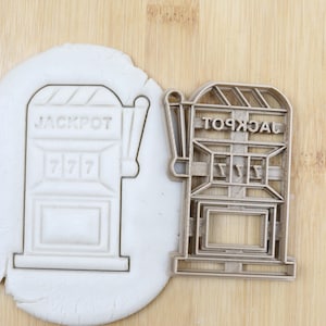 Könnte beinhalten: Ein silberfarbener Metall-Ausstecher in Form eines Spielautomaten mit dem Wort "JACKPOT" oben und "7 7 7" auf der Vorderseite. Der Ausstecher befindet sich auf einer Holzoberfläche neben einem weißen Teigabdruck des Ausstechers.