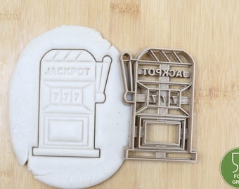 May include: A silver-colored metal cookie cutter shaped like a slot machine with the word "JACKPOT" on the top and "7 7 7" on the front. The cookie cutter is on a wooden surface next to a white dough imprint of the cookie cutter.