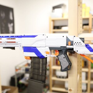 May include: A white and blue toy gun with an orange stock and a black magazine. The gun is mounted on a wooden shelf.