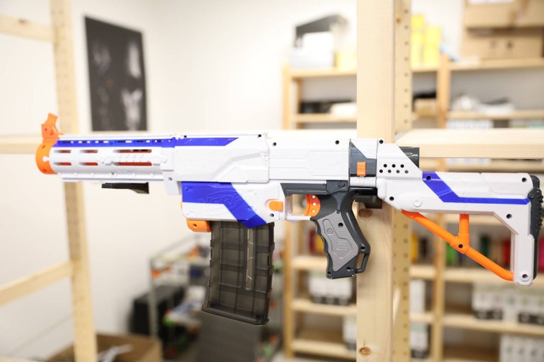 May include: A white and blue toy gun with an orange stock and a black magazine. The gun is mounted on a wooden shelf.