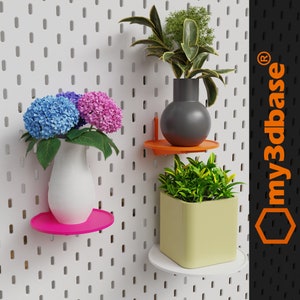 Puede incluir: Tres plantas en macetas de diferentes colores se muestran en estantes blancos unidos a una pared de tablero de clavijas blanco. Los estantes son naranja, rosa y blanco. Las plantas son una planta verde en una maceta amarilla, una planta verde en una maceta gris y un ramo de flores rosas y azules en un jarrón blanco.