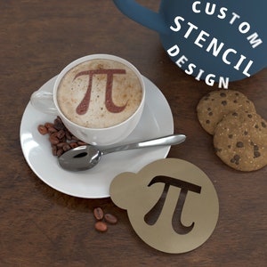 Könnte beinhalten: Eine braune und goldene Schablone mit dem Symbol Pi ausgeschnitten. Die Schablone liegt auf einer Holzoberfläche neben einer Tasse Kaffee mit einem Pi-Symbol im Schaum. Auf der Oberfläche befinden sich auch zwei Schokoladenkekse.