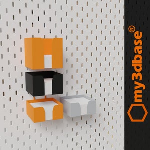 Könnte beinhalten: Drei Wandhalterungen für Notizblöcke. Die Halterungen sind aus Kunststoff und in den Farben Orange, Schwarz und Grau erhältlich. Die Halterungen sind an einer weißen Lochwand befestigt, auf der im oberen rechten Eck der Text "my3dbase®" steht.